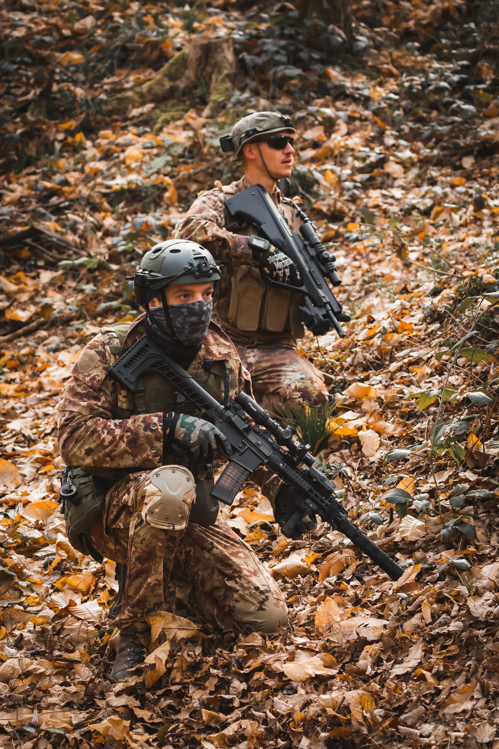 Two X-Tag players in woodland camouflage advancing on one knee through an autumn forest